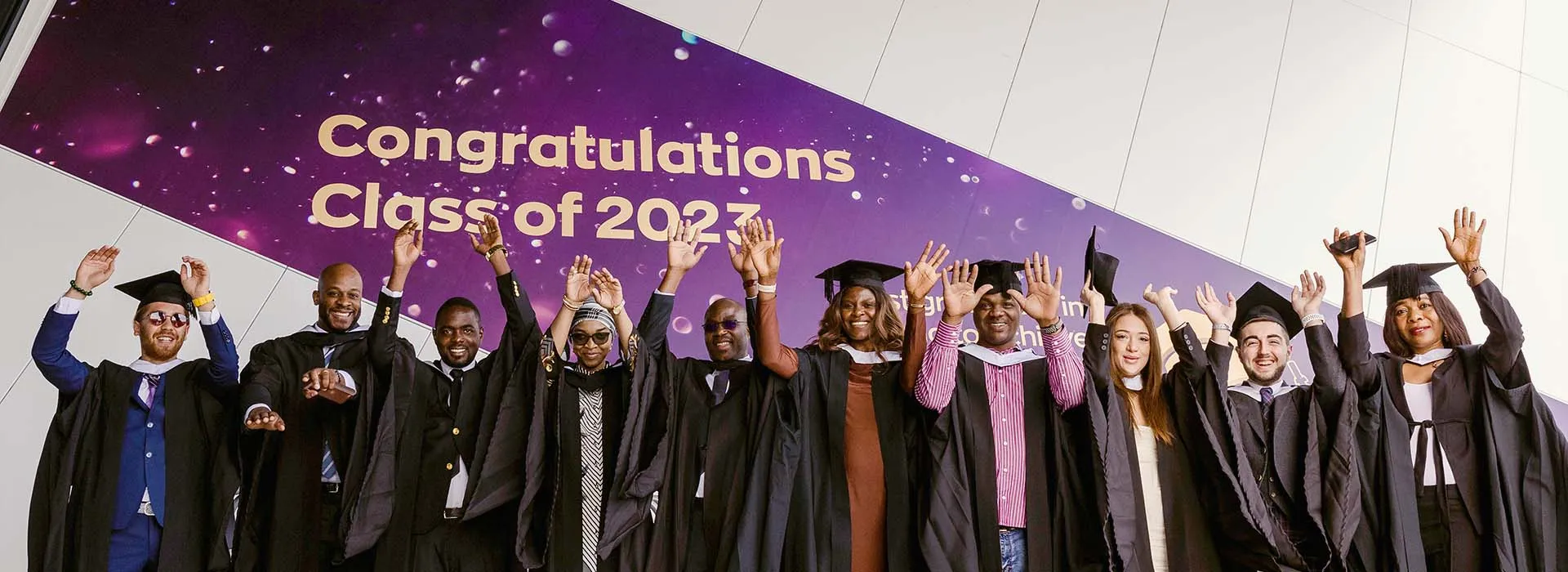 Graduates in gowns and hats with their hands in the air. Behind them is a sign saying 'Congratulations Class of 2023'