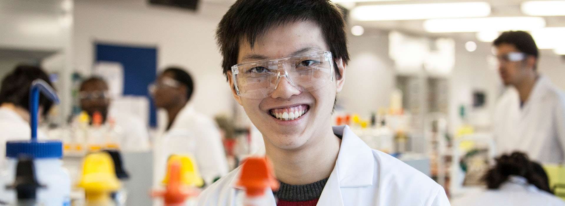 student in a lab coat and safety glasses smiling in front of science equipment