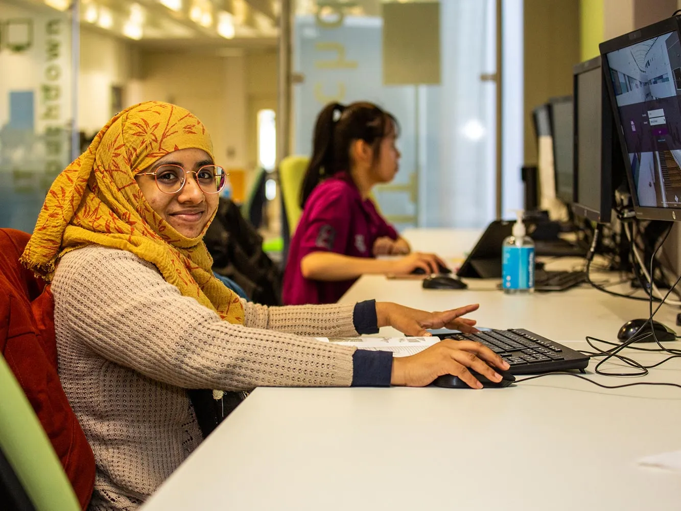 student wearing a headscarf sitting at a computer desk smiling