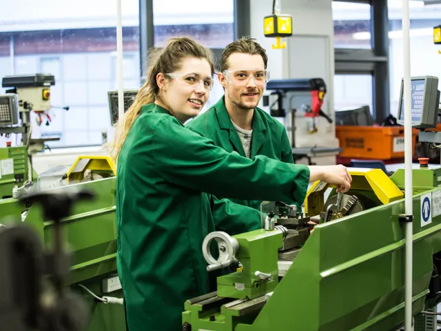 two students in engineering overalls at machines