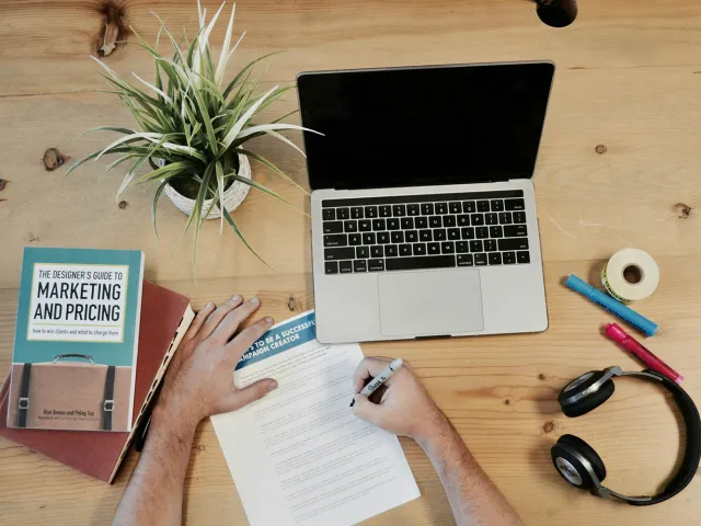 person writing at a desk with a book, laptop, headphones
